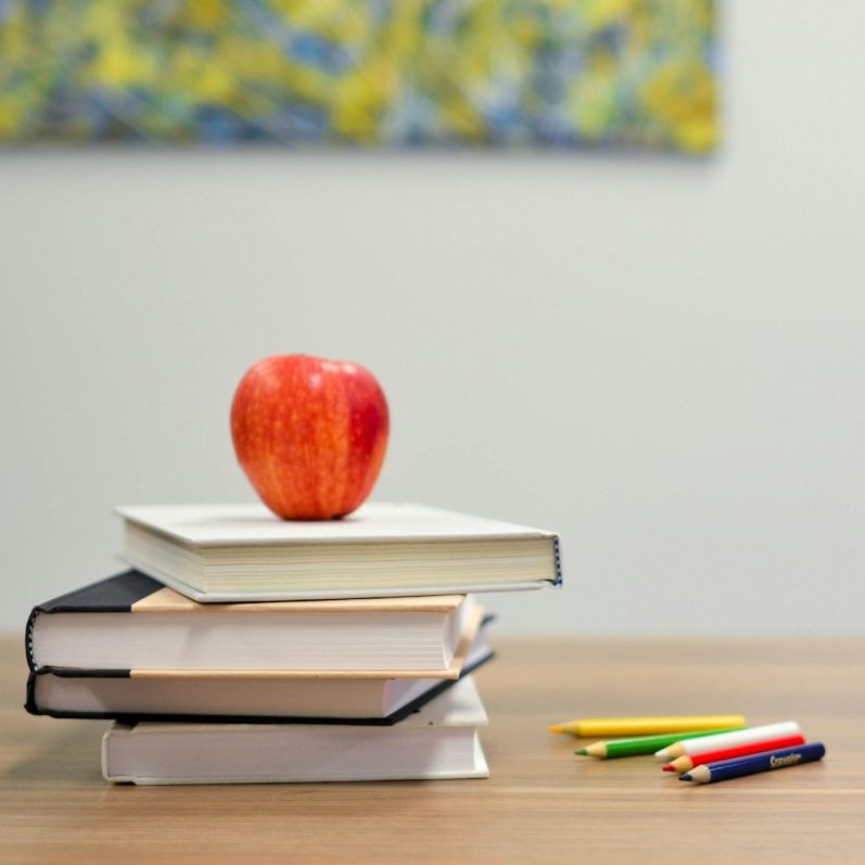 Klassenzimmer Roter Apfel auf einem Stapel Bücher, daneben bunte Stifte. Stilisierte Szene aus einem Klassenzimmer.